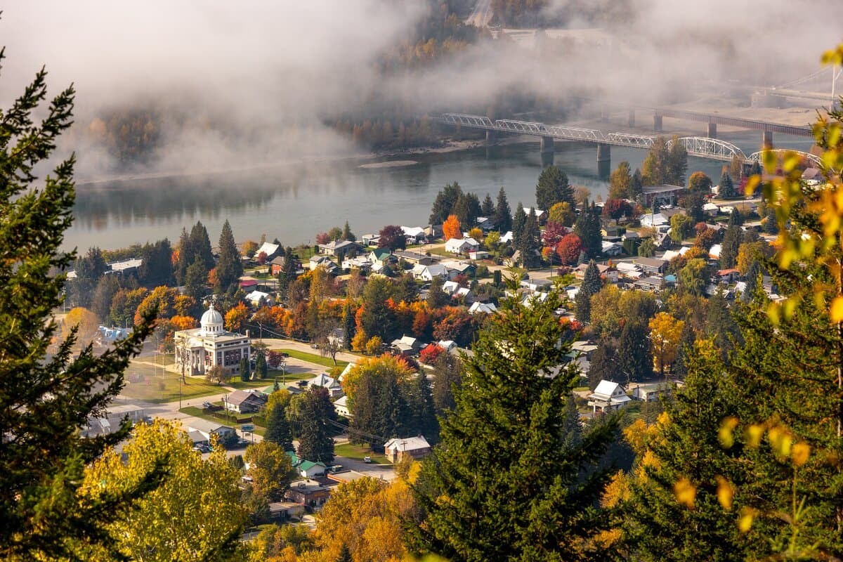 Aerial view of a town with colorful autumn trees, a river with a bridge, and misty hills in the background.