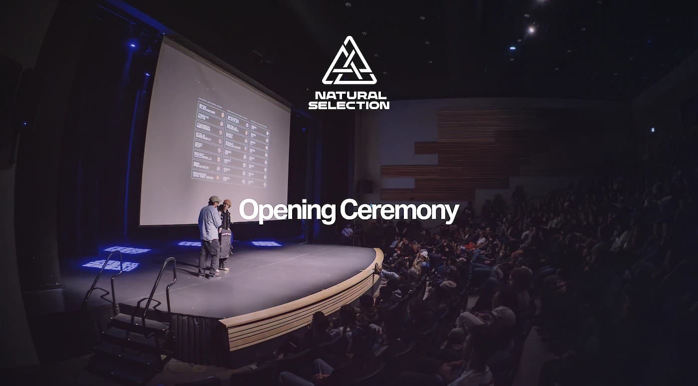 Wide-angle view of a stage with two speakers, large screen, and audience. "Natural Selection Opening Ceremony" is projected on the screen.