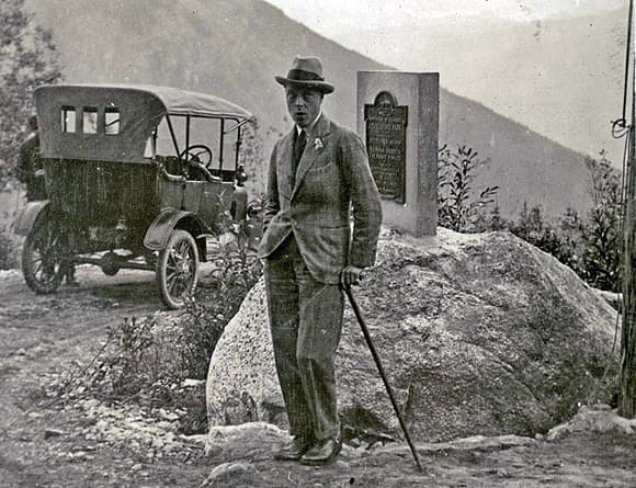 A man in a suit and hat, holding a cane, stands by a rock with a plaque. An early 20th-century car is parked nearby, with mountains in the background.