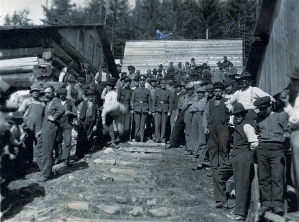 A large group of men in early 20th-century attire gather on wooden steps between buildings, with trees in the background.