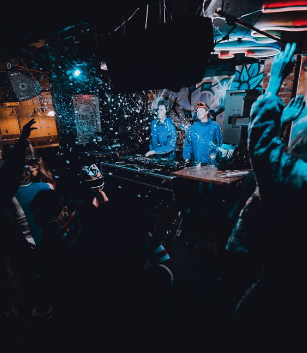 Two DJs in blue hoodies play amid confetti and neon lights as a crowd raises hands in a graffiti-filled club