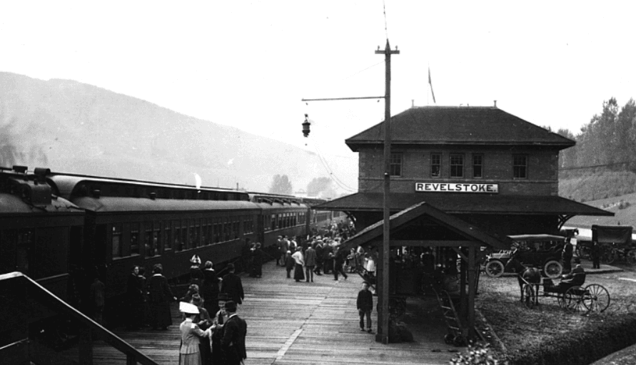 Historic train station scene with a steam train, people on the platform, and horse-drawn carriages. Sign reads "Revelstoke."
