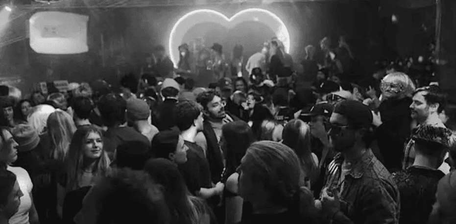 A crowded club party in black and white features people dancing under dim lighting, with a large illuminated heart in the background.