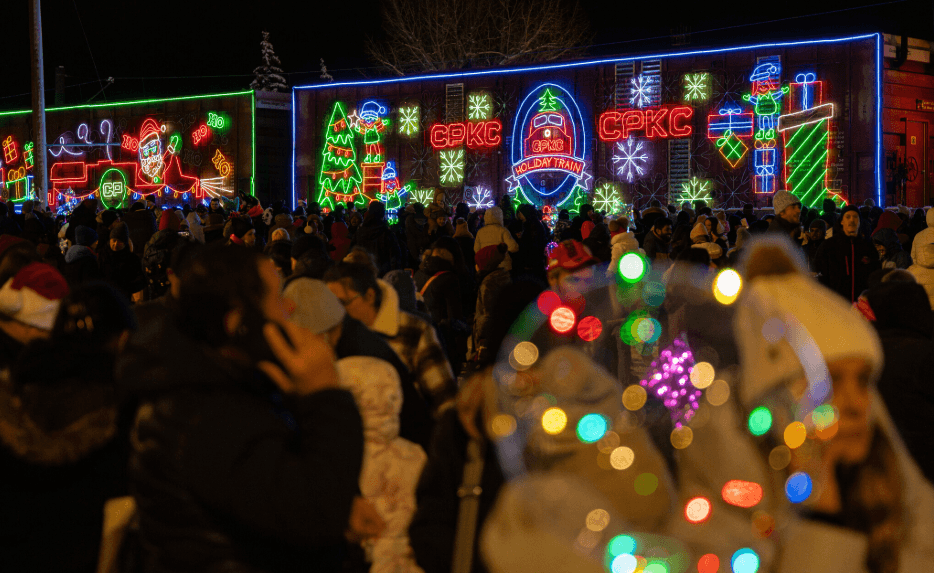 Crowd gathered at night in front of a brightly lit train with colorful holiday-themed neon decorations and snowflake designs.