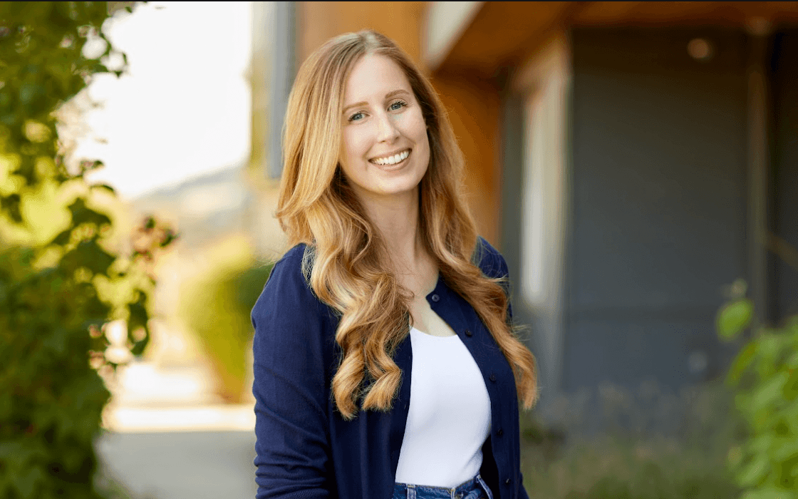 Kyla Egan - Tourism Revelstoke Executive Director A woman with long, wavy blonde hair, wearing a navy cardigan and white top, smiles outdoors with greenery and a building in the background.
