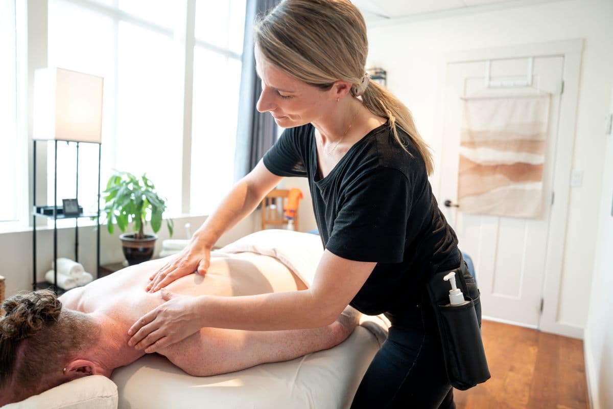 Person receiving a back massage in a bright room with a plant and a towel on the wall. The masseuse is wearing a black shirt.