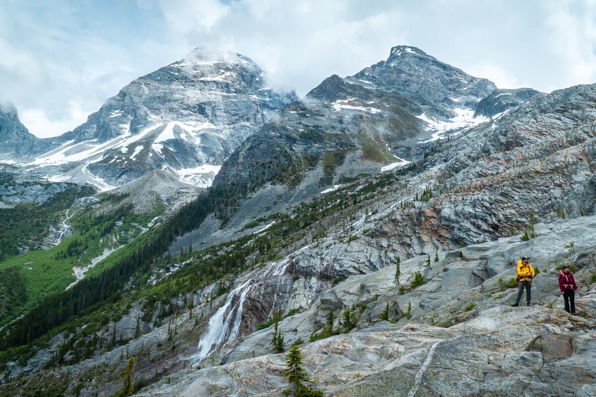 Two hikers in colorful jackets traverse a rocky mountain landscape with snow-capped peaks and a waterfall under a cloudy sky.