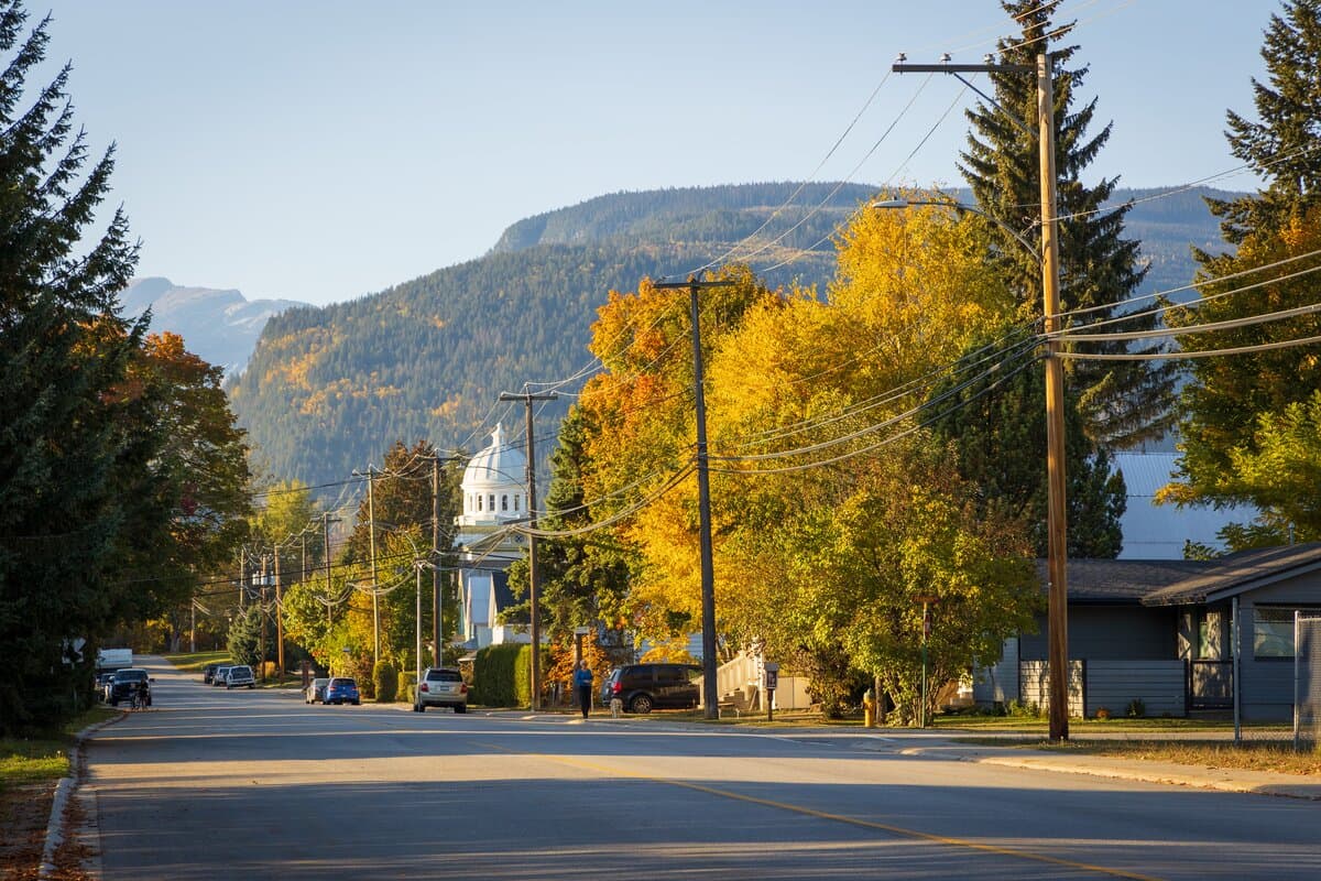 A quiet street lined with colorful autumn trees, power lines, and houses, with mountains and a domed building in the background.