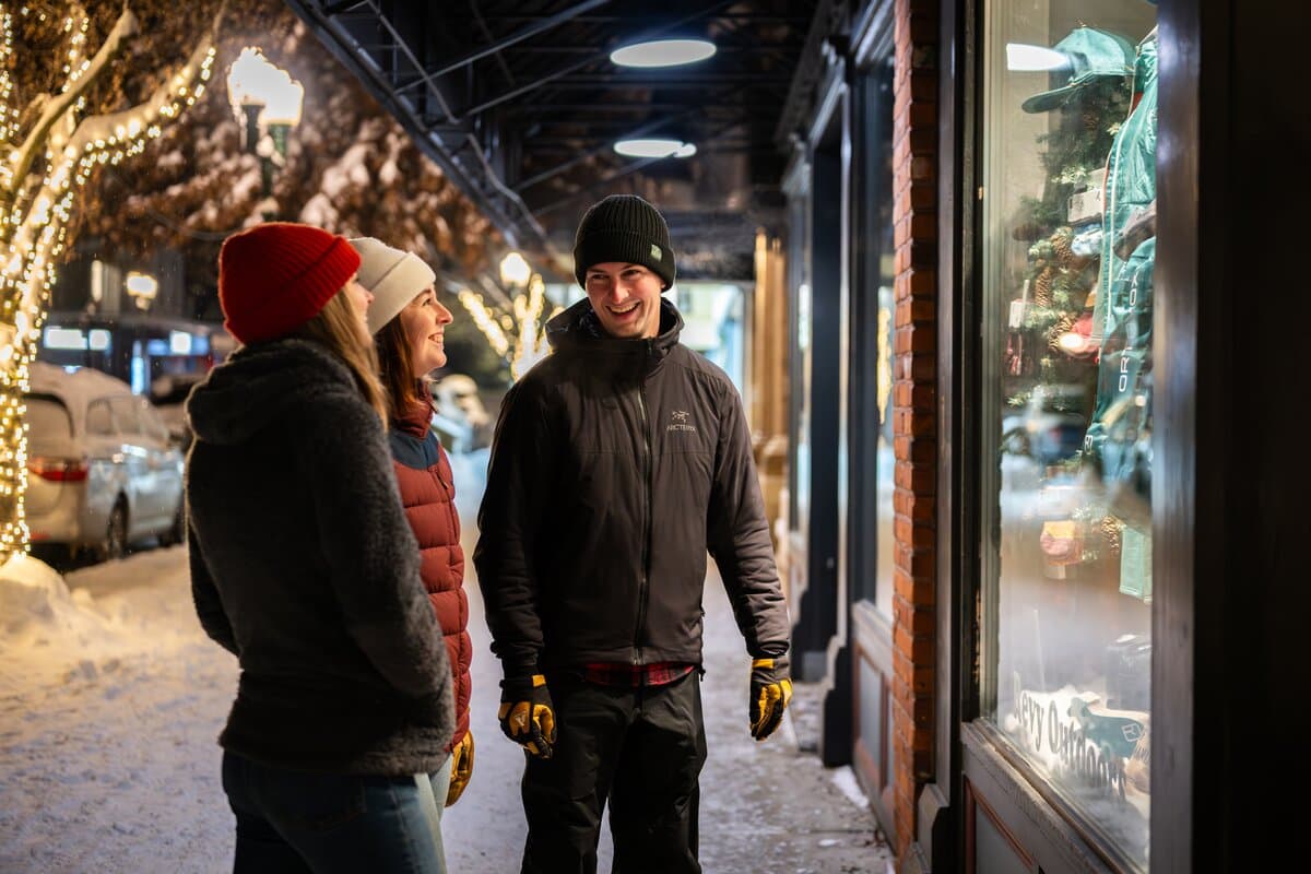 Three people in winter clothing smile while window shopping on a snowy, decorated street at night.
