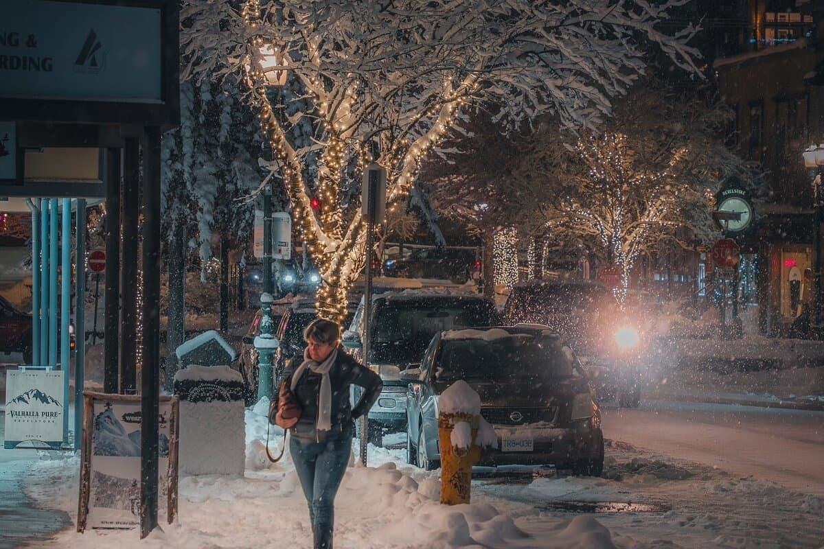 Person walking on a snowy street at night, with trees wrapped in string lights and cars parked along the road.