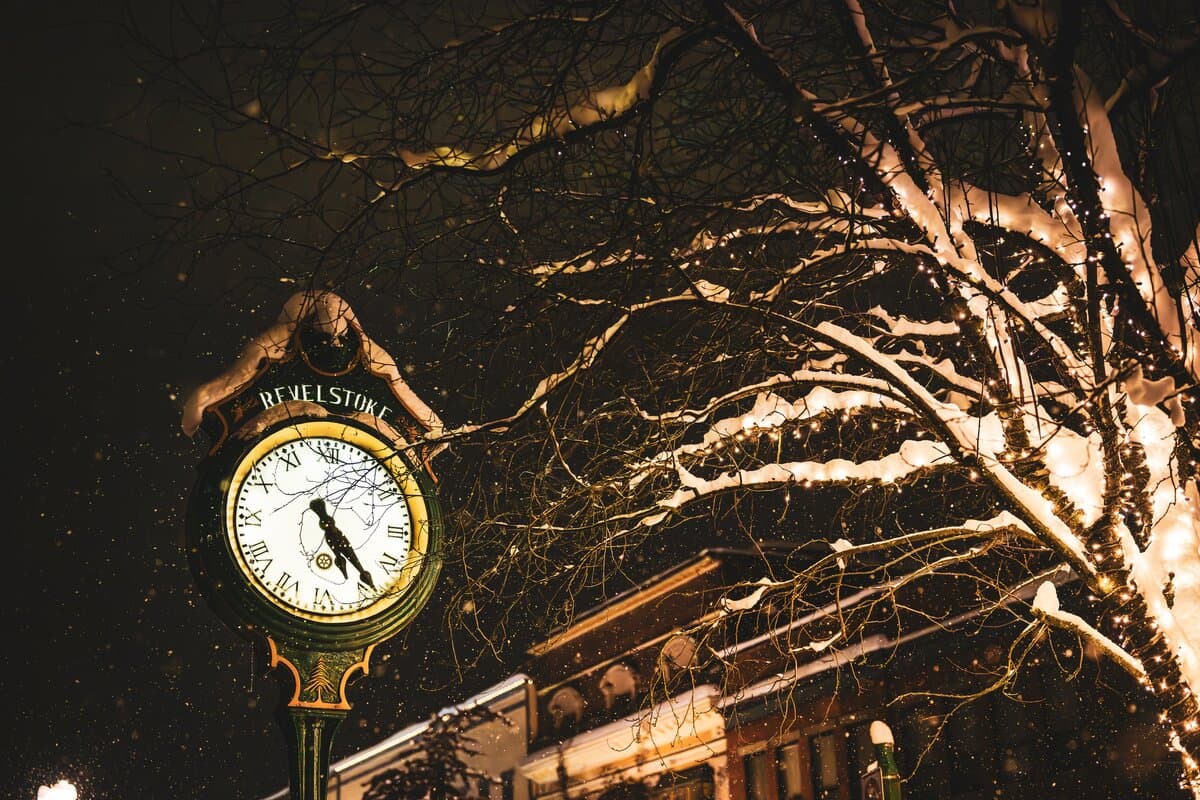 A snow-covered clock shows 9:15 beside a tree with twinkling lights on a snowy night in a town square.