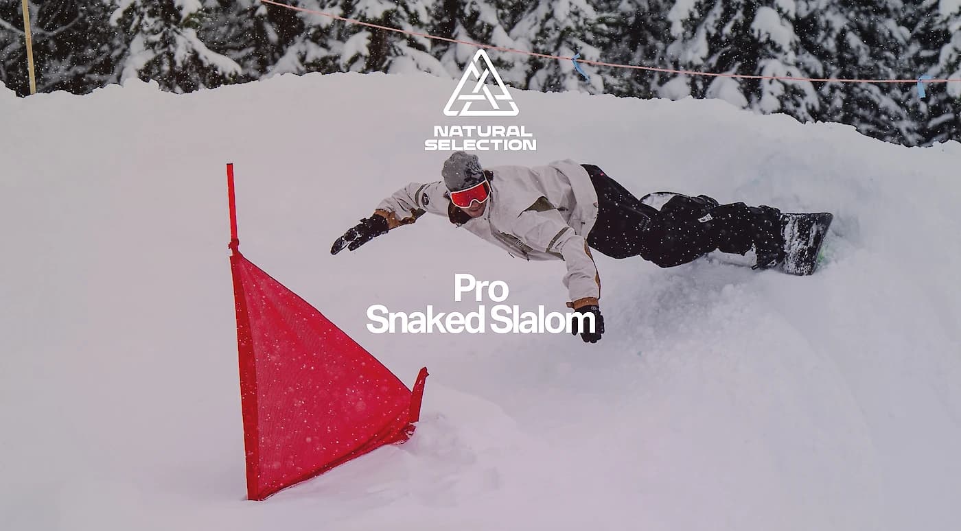 Snowboarder in mid-air during a slalom, wearing a white jacket and black pants with snow-covered trees in the background.
