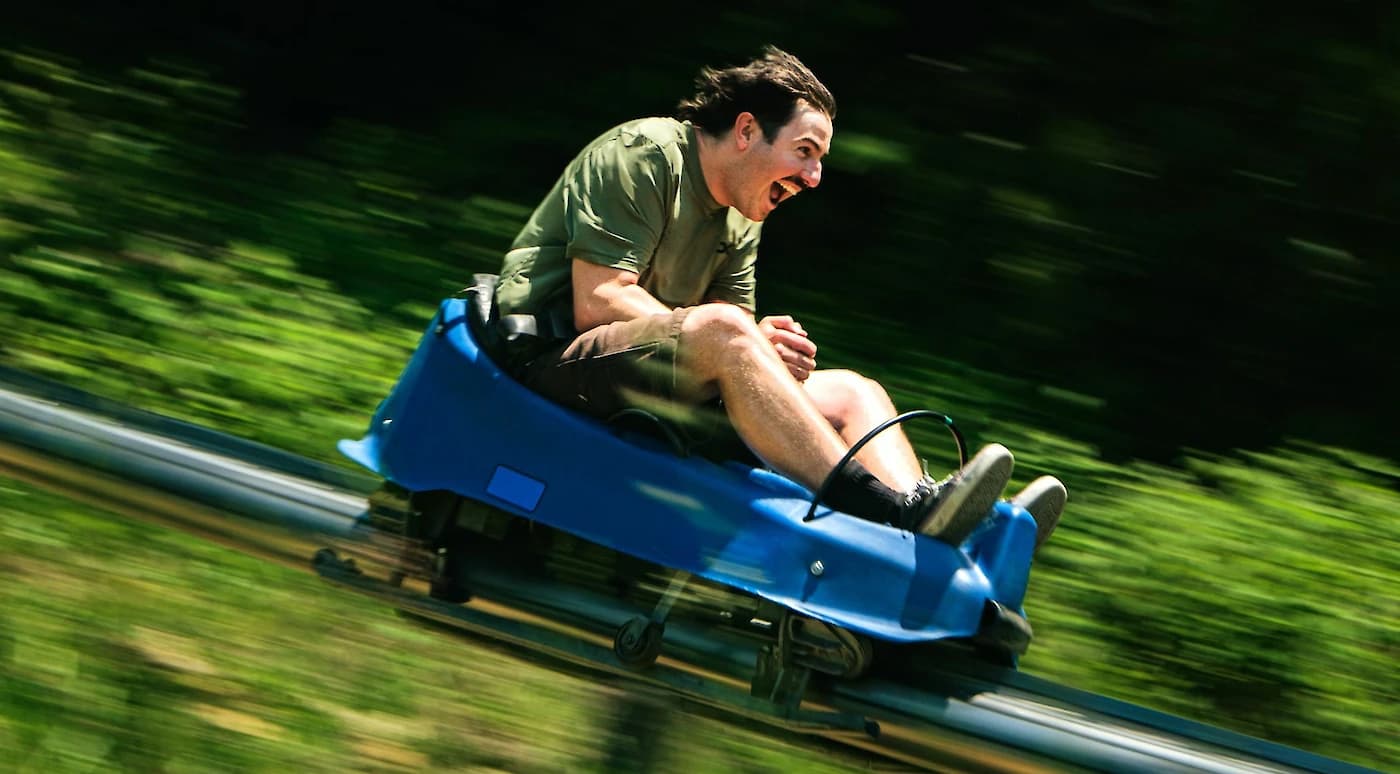 Man joyfully riding a blue alpine coaster, moving swiftly with blurred greenery in the background.