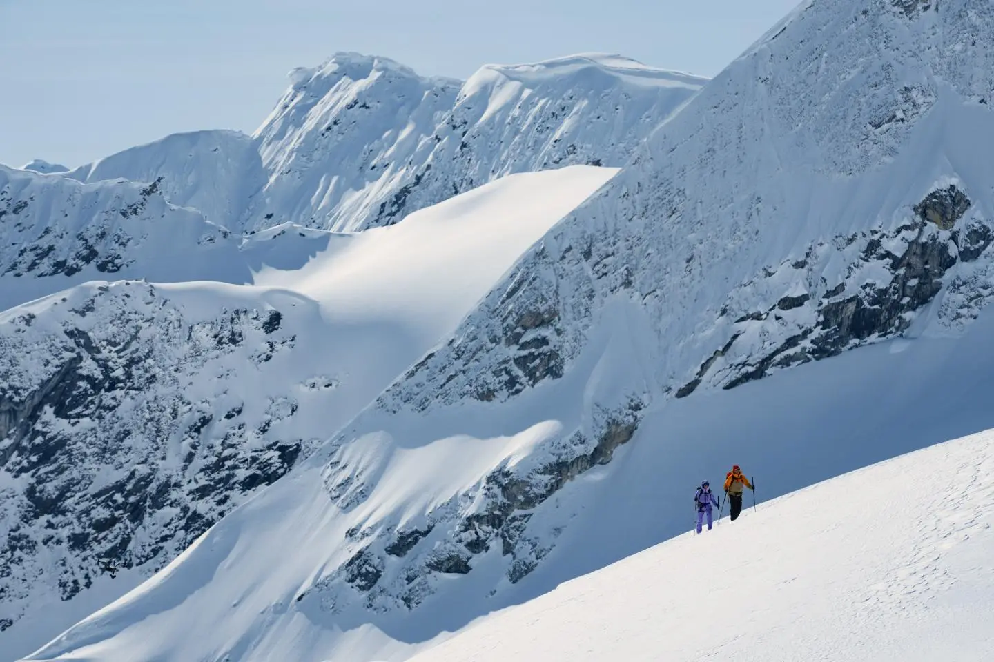 Two people hiking up a snowy mountain slope with jagged peaks in the background under a clear blue sky.
