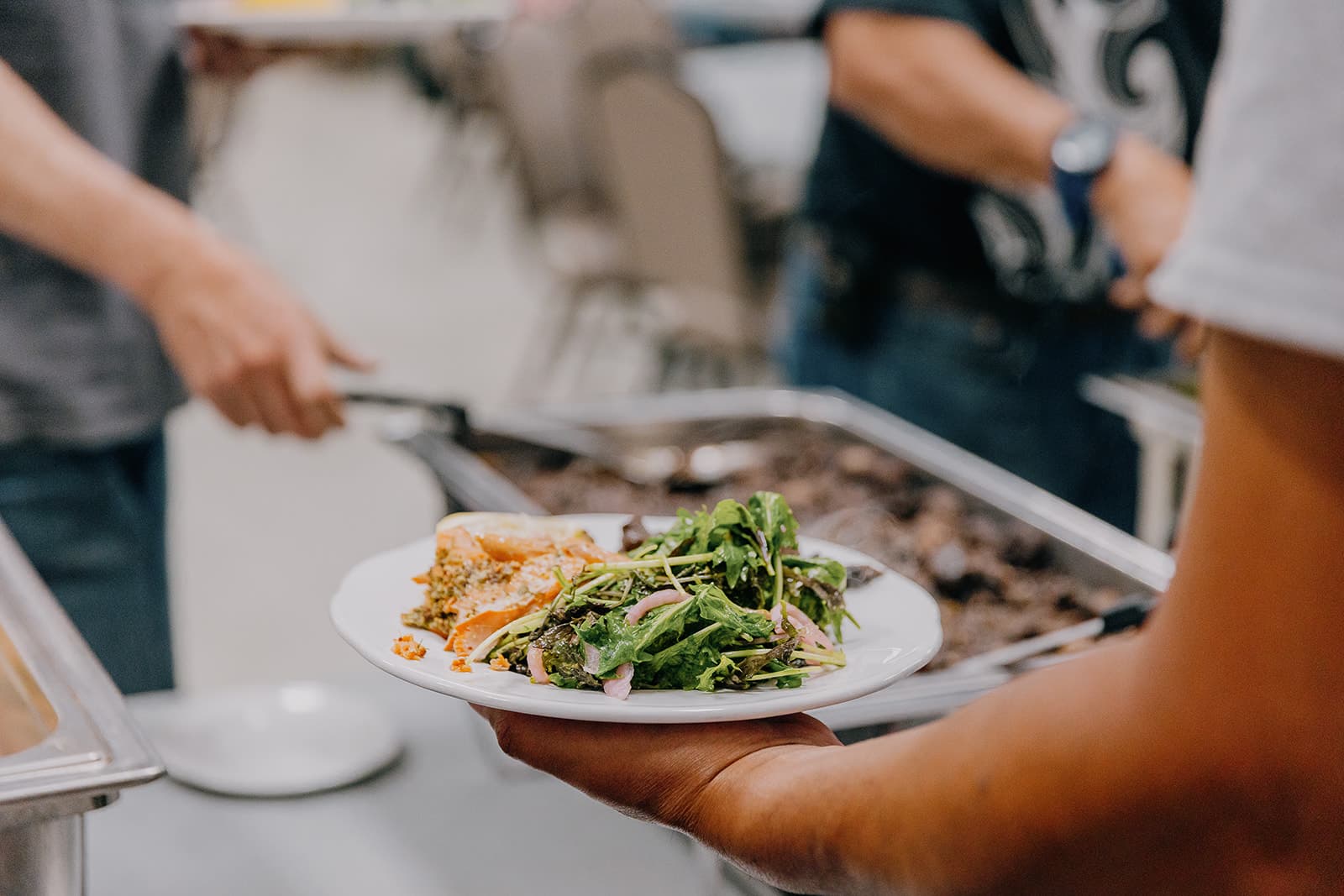 Person holding a plate with salad and lasagna, standing in a buffet line where another person is serving food from a tray.