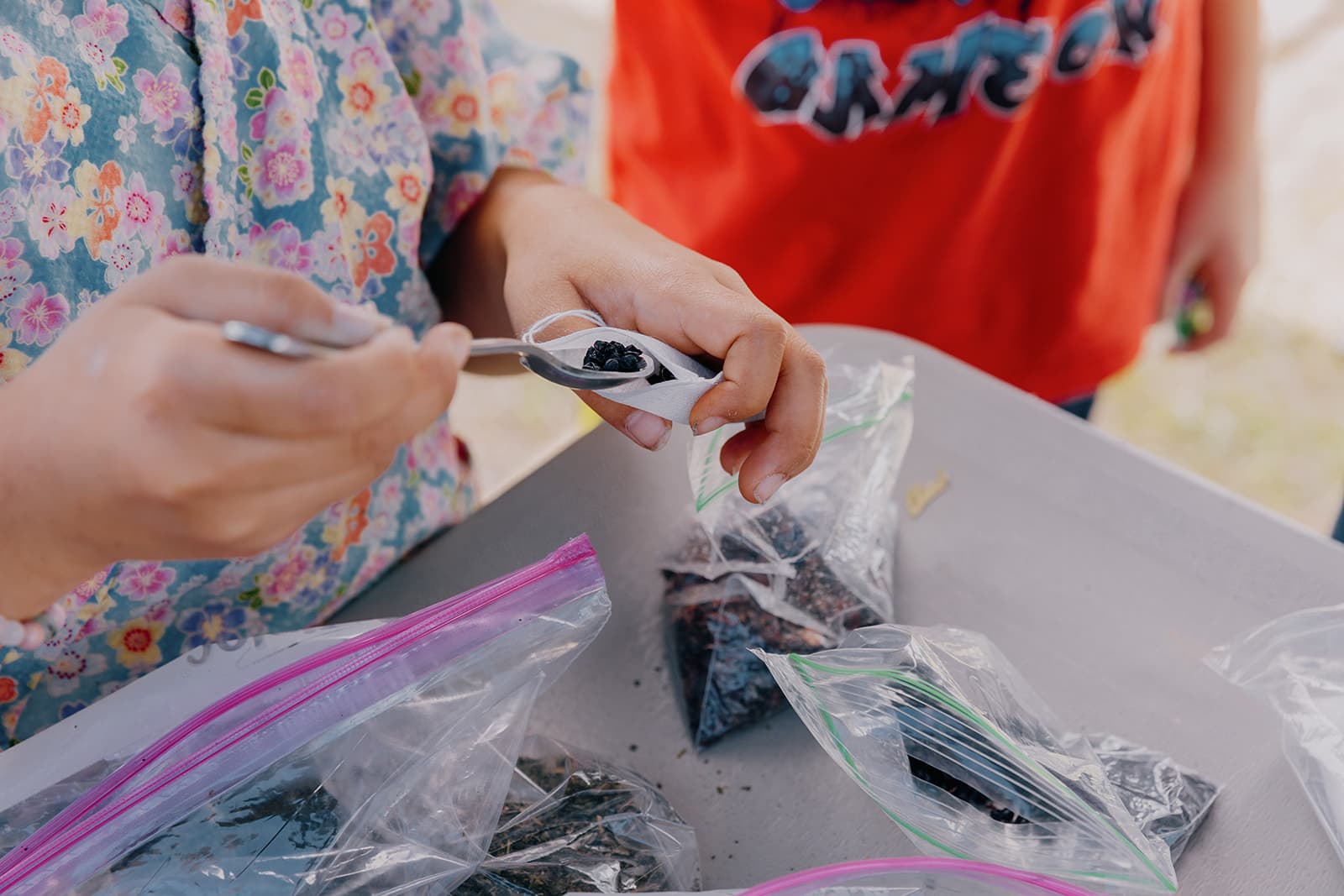 Person scooping black seeds into a small bag on a table with other bags. They are wearing a floral shirt.