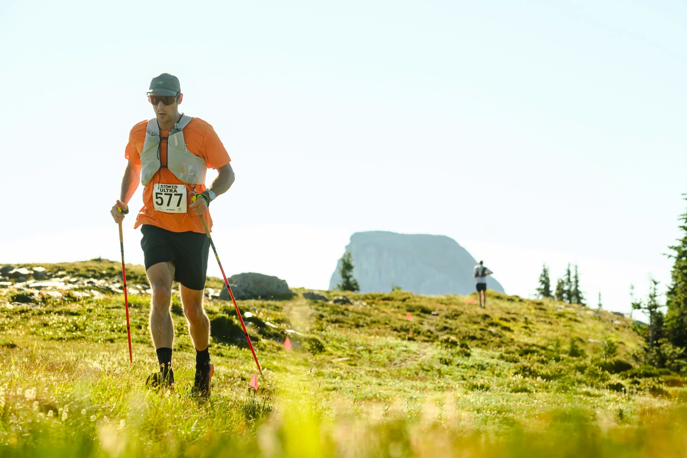 Trail runner in an orange shirt and cap, using poles, traverses a grassy mountain landscape with a distant rock formation in view.