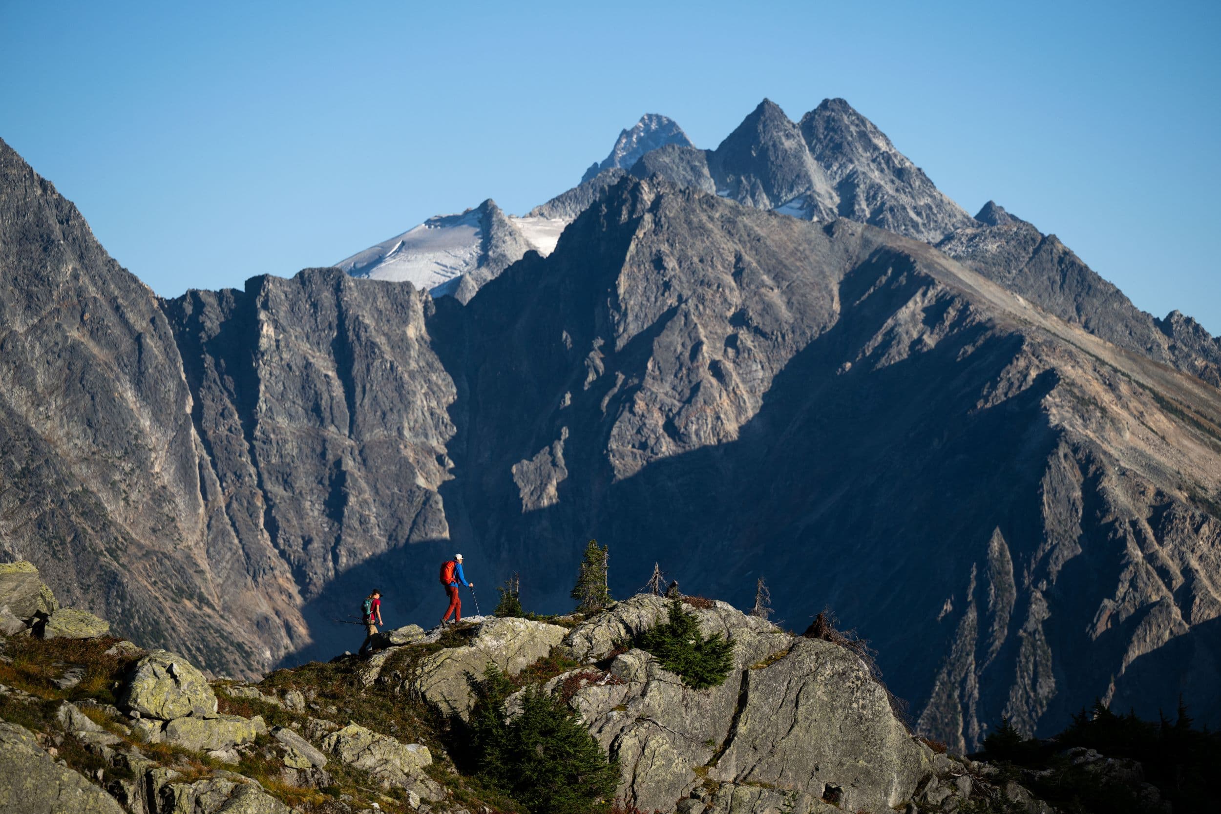 Two hikers in red jackets stand on a rocky ridge with towering, jagged mountains in the background under a clear blue sky.