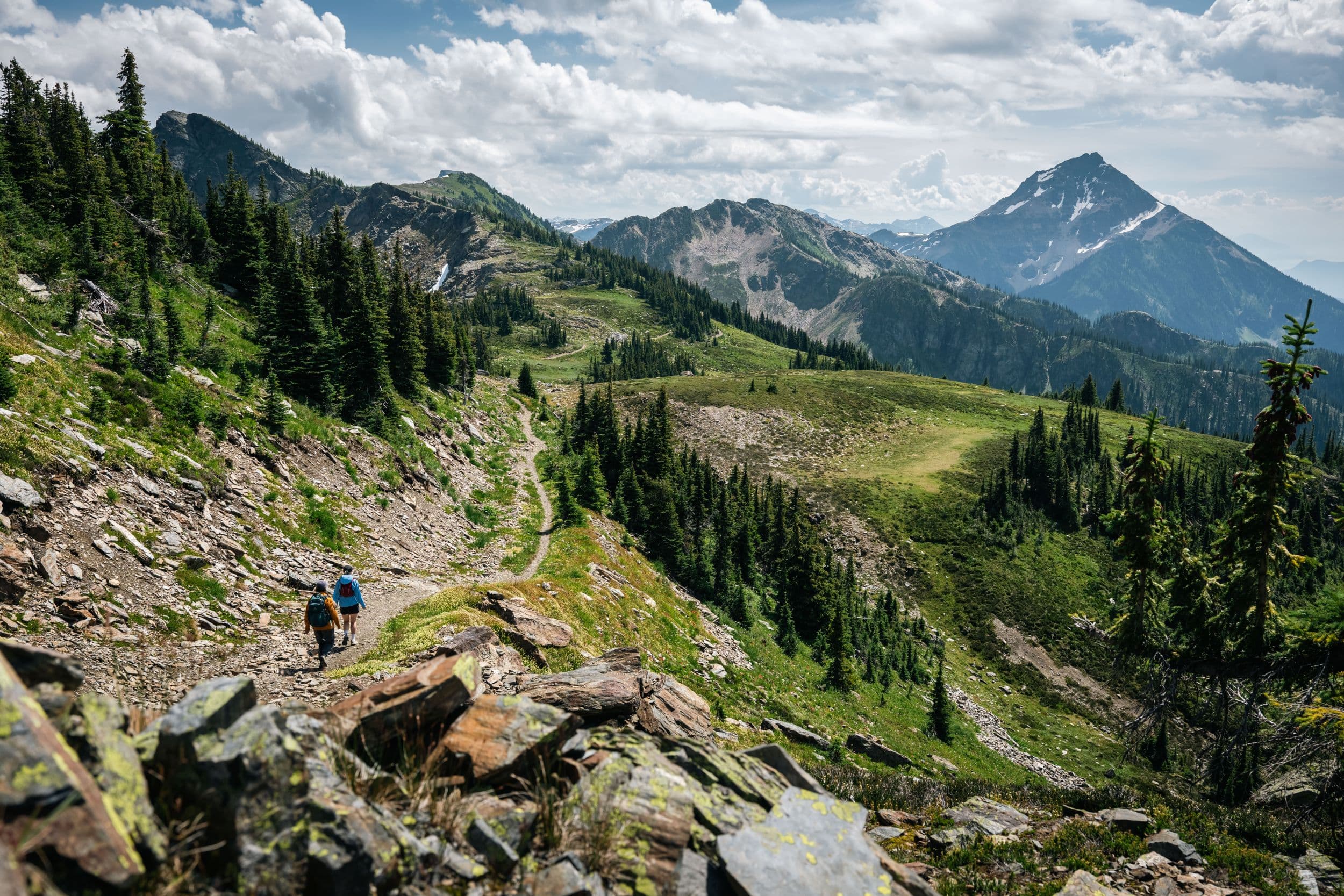 Two hikers on a rocky alpine trail winding through green ridges and fir trees toward a distant snow-capped mountain under a cloudy sky.