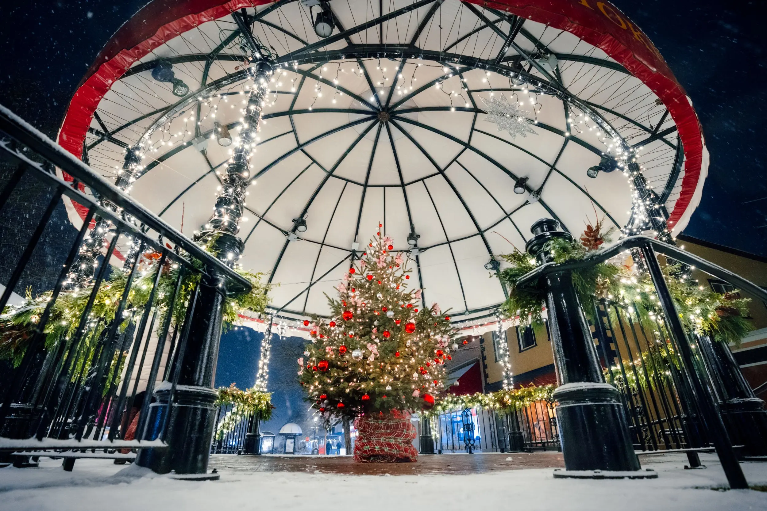 Snow-dusted bandstand lit with white string lights, garlands, and a decorated Christmas tree with red ornaments.
