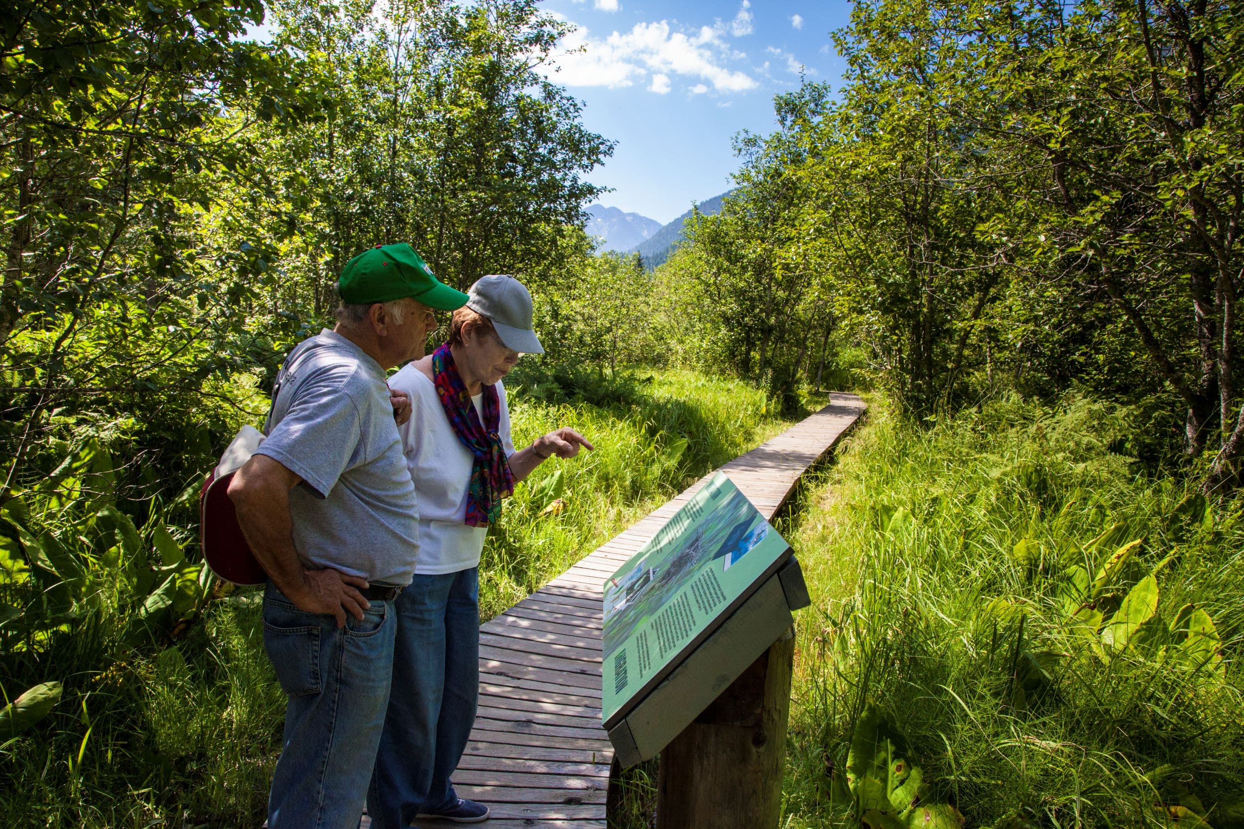 Two people study an informational sign on a wooden path surrounded by lush greenery and trees under a clear blue sky.