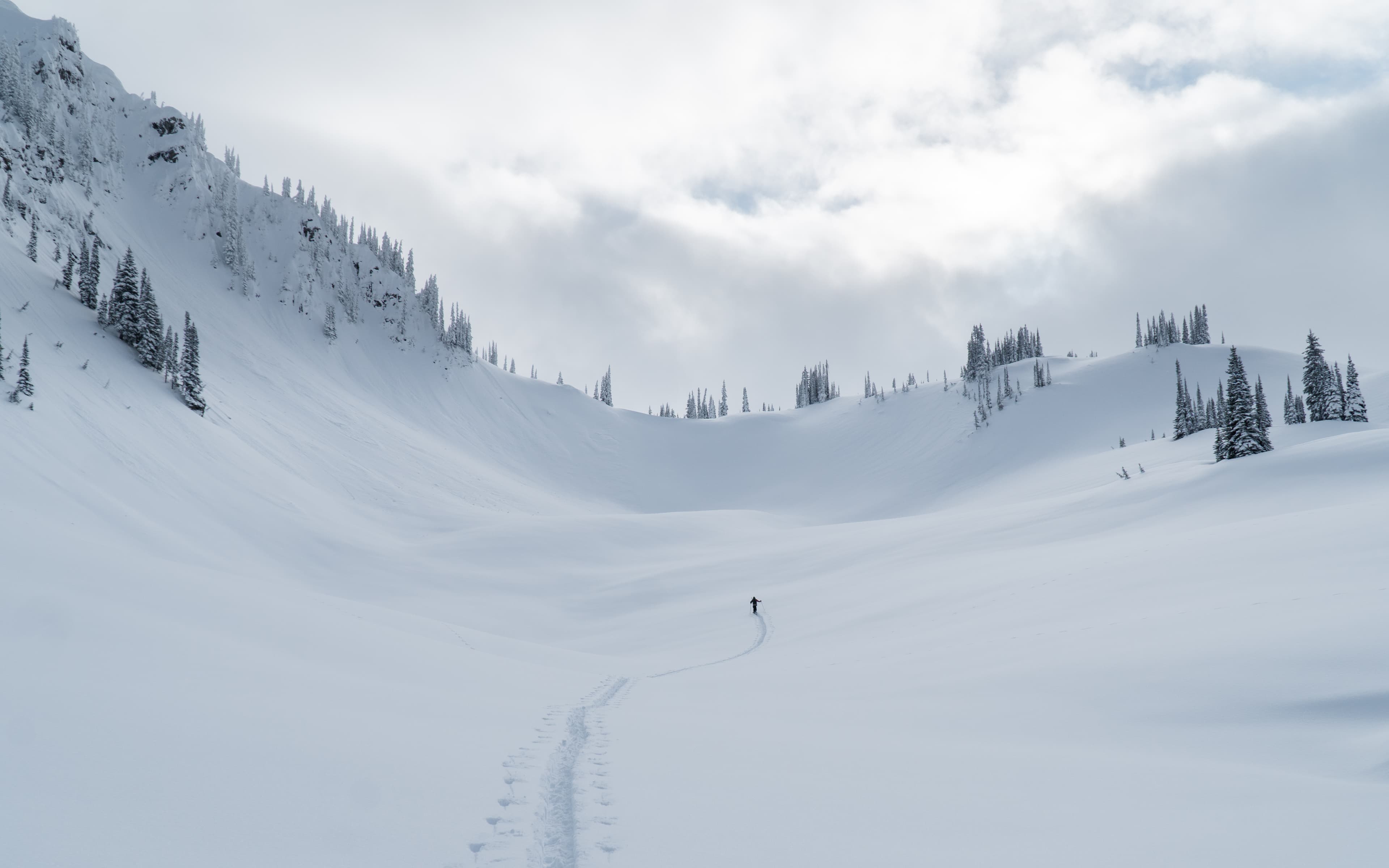 A lone skier traverses a vast, snow-covered mountain landscape under a cloudy sky, leaving a single trail behind.