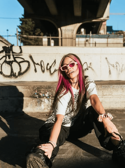 Woman with pink hair streaks and sunglasses sits on the ground, smiling, in front of graffiti-covered concrete under a bridge.