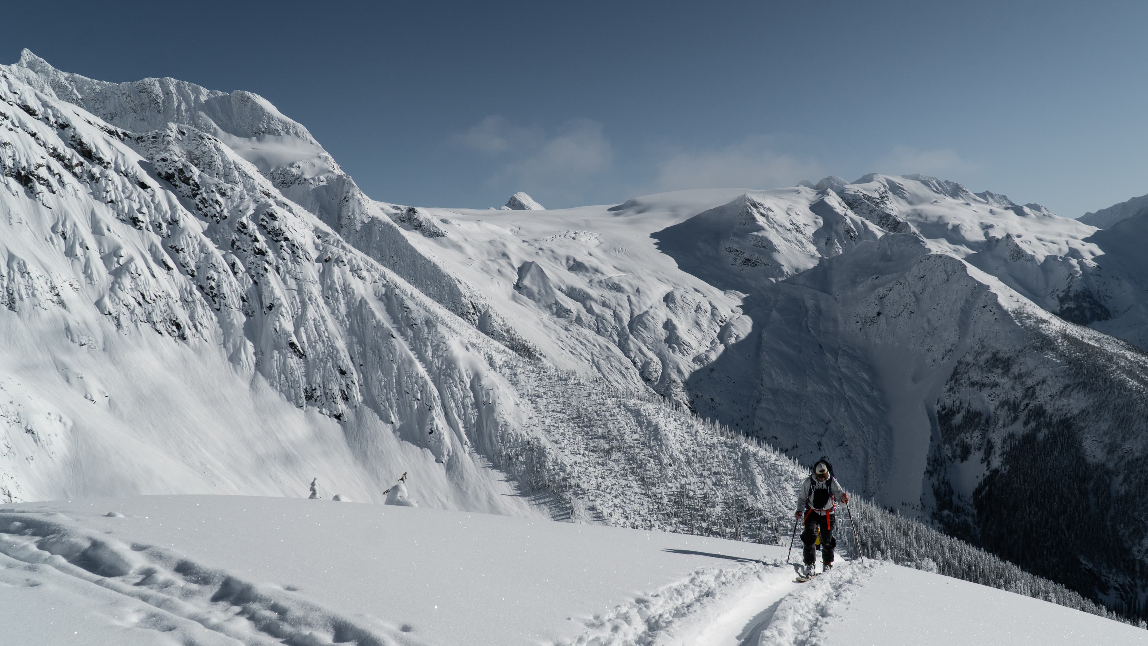 A person with a backpack hikes up a snowy mountain trail, surrounded by vast snow-covered peaks under a clear blue sky.