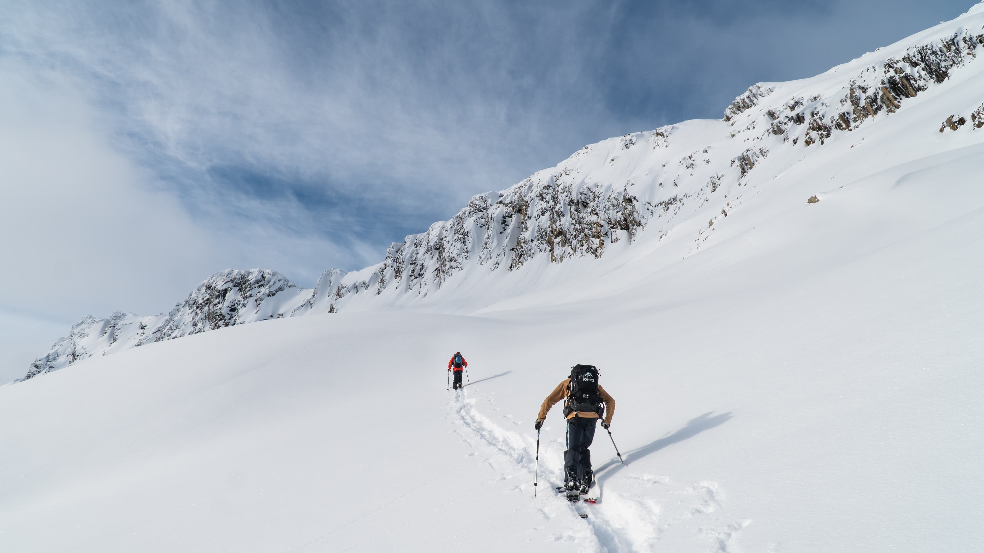 Two skiers ascend a snowy mountain slope under a partly cloudy sky, leaving tracks behind them. Rocky peaks are visible in the background.
