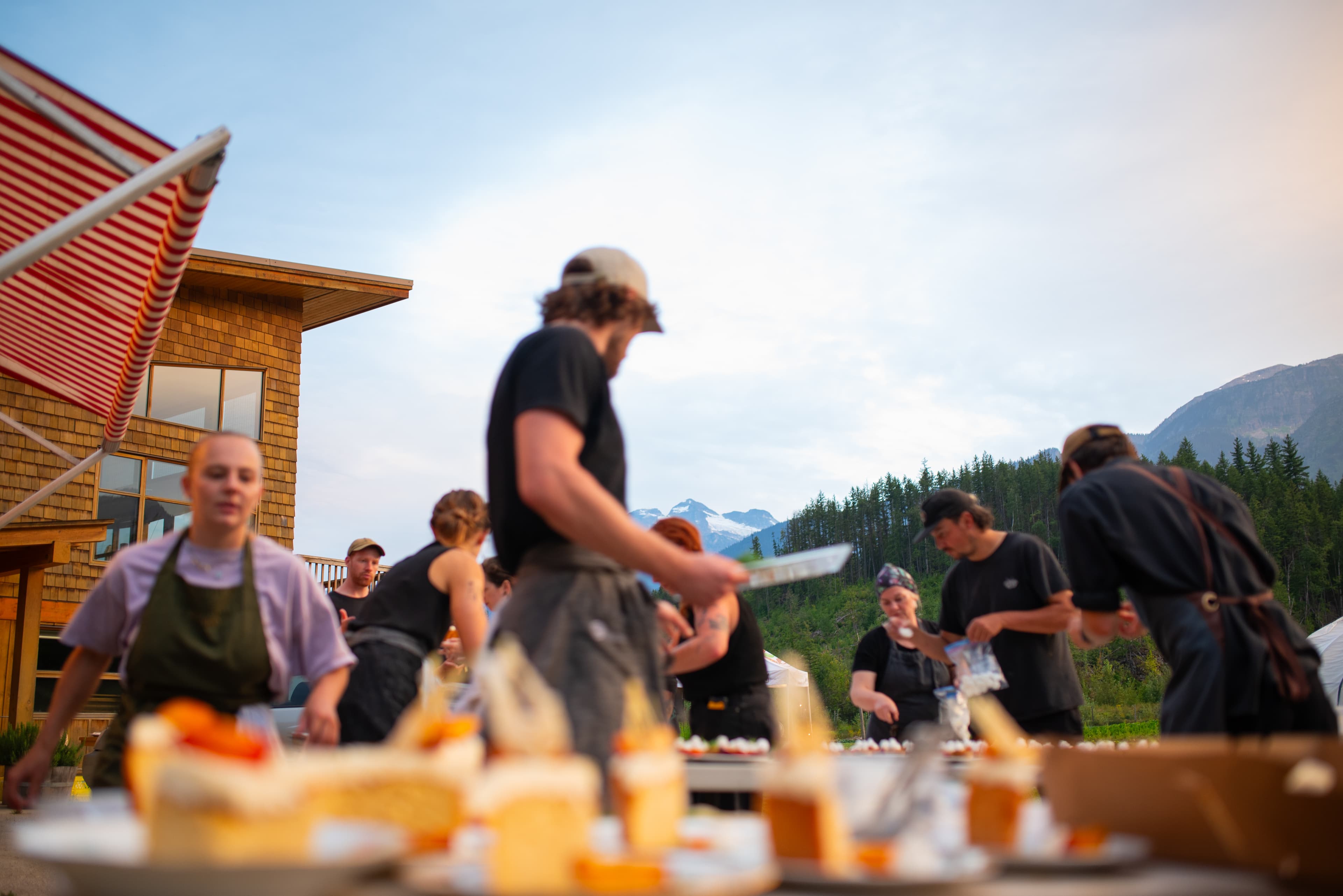 People preparing food outdoors with a mountain view in the background, under a striped awning near a wooden building.