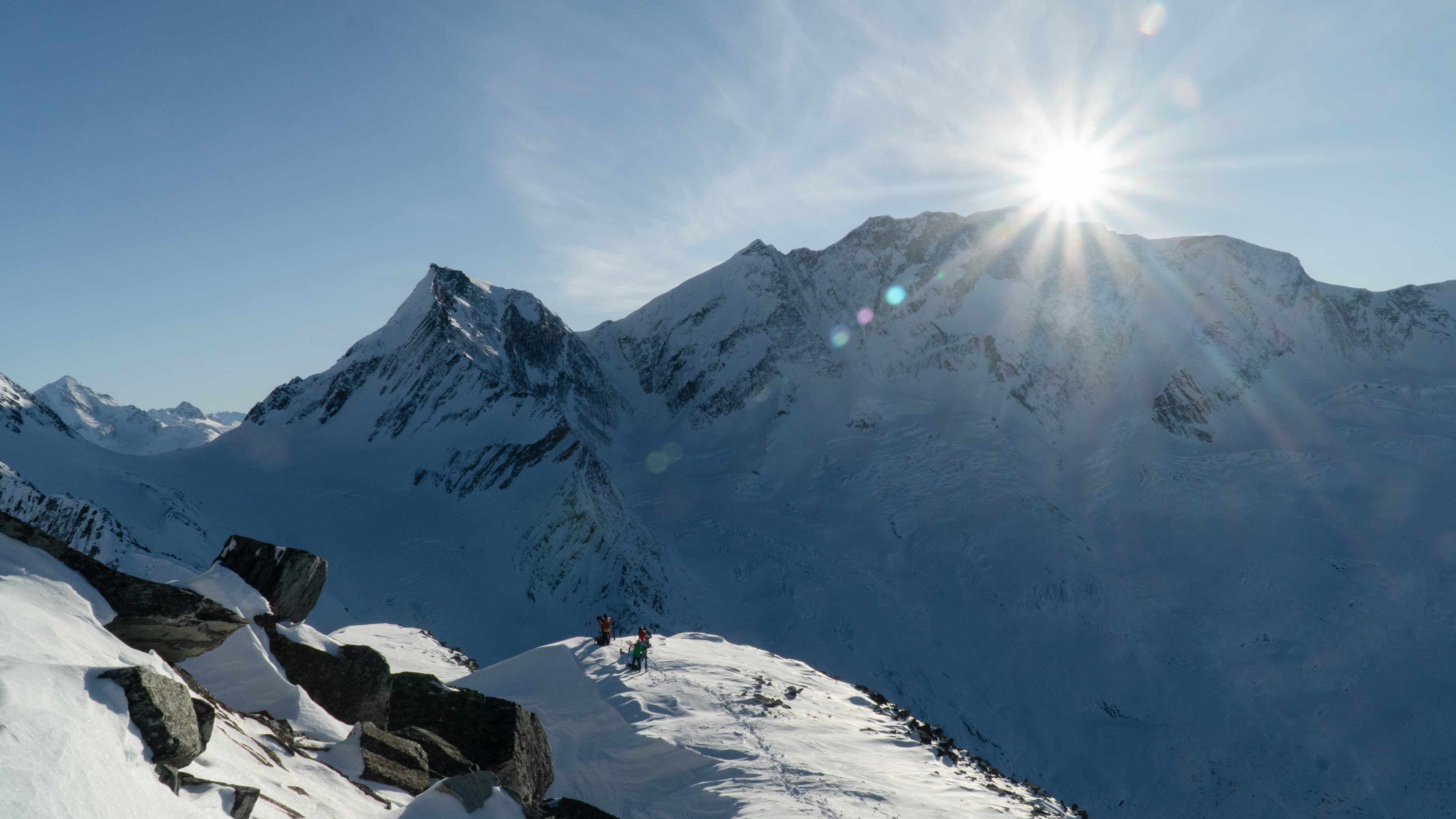Snowy mountain landscape with two climbers on a ridge, sun shining brightly over distant peaks under a clear blue sky.