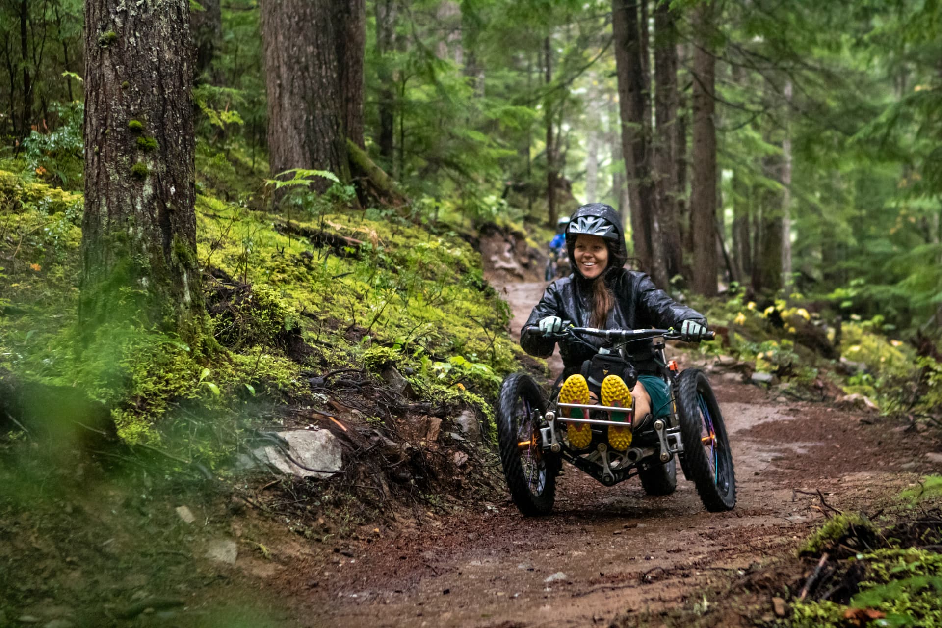 an adaptive biker smiles as she rolls down a wet trail