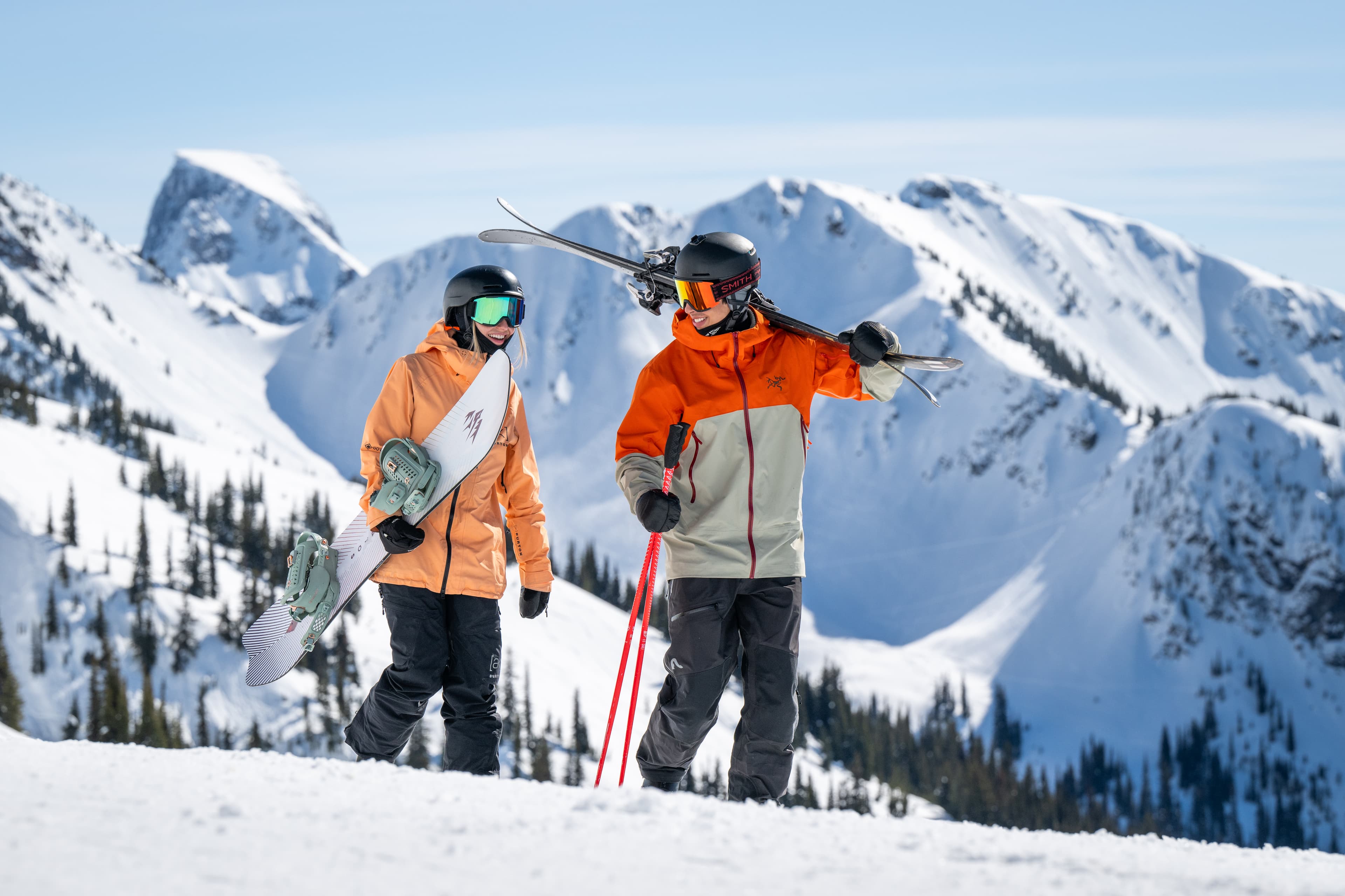male and female holding skis and snowboard on mountain ridge