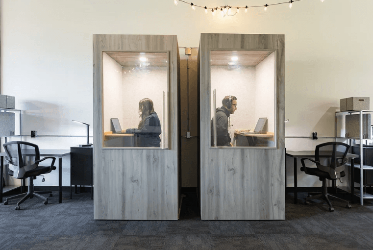 Two people working in adjacent, soundproof booths with laptops, separated by a wall. Office chairs and shelves are visible in the background.
