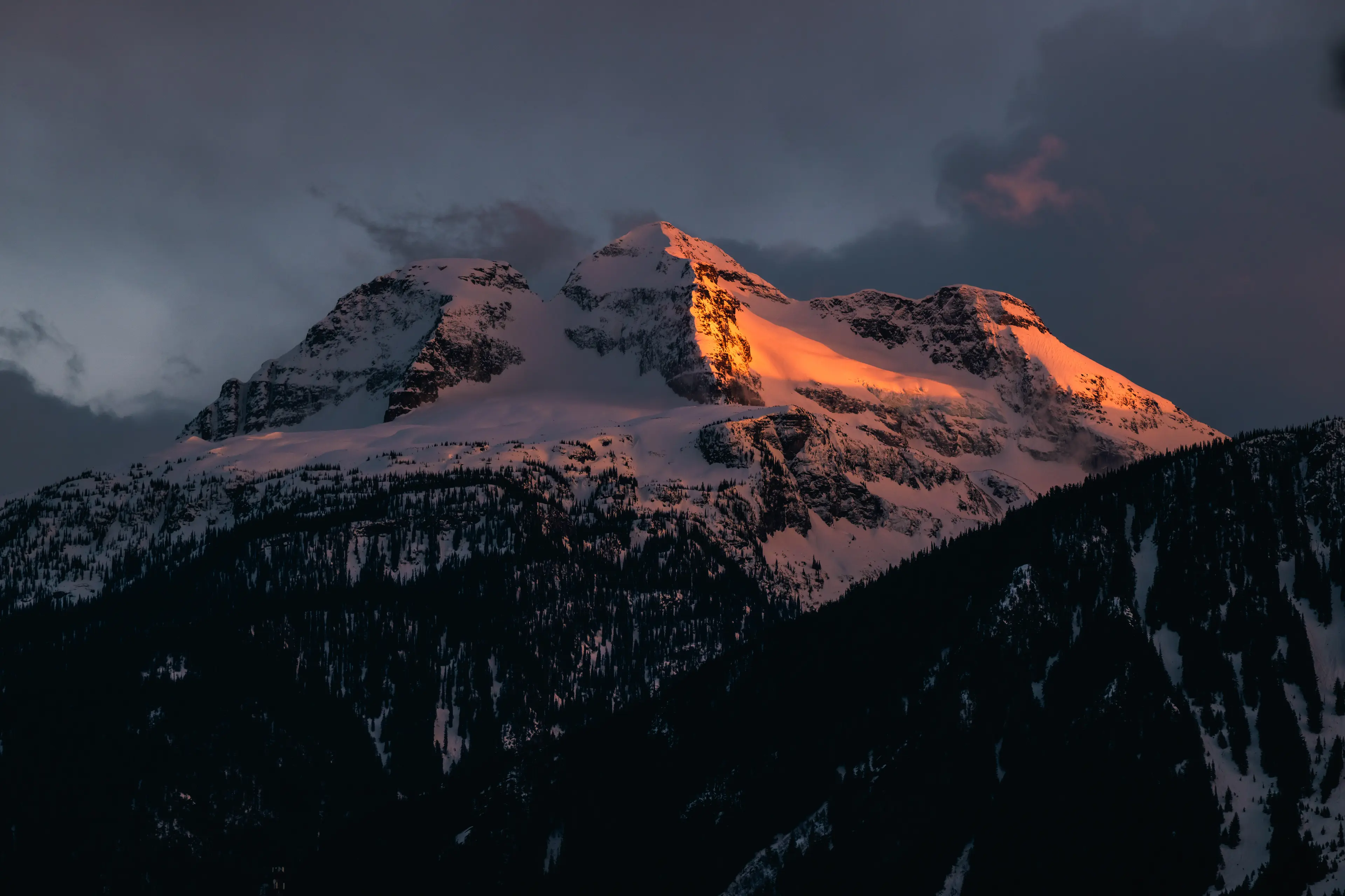 Snow-covered mountain peaks bathed in orange alpenglow at sunset, looming above dark forested slopes under a moody twilight sky.